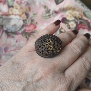 May include: A ring featuring a large, circular, porous focal piece with a dark brown and metallic gold finish. The surface of the focal piece is covered in numerous small, irregular indentations, creating a textured, crater-like appearance.