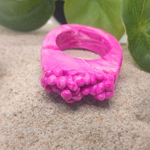 May include: A bright pink ring with a marbled finish and a cluster of small, rounded, bubble-like protrusions on the front surface. The band is thick and smooth, featuring a glossy texture that highlights the swirling shades of pink throughout.