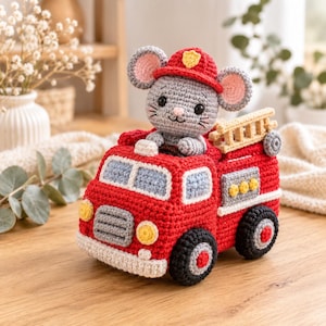 Op de afbeelding: Een gehaakte grijze muis met een rode brandweerhelm, zittend in een felrode gehaakte brandweerwagen. De brandweerwagen is voorzien van een lichtgekleurde ladder aan de achterkant, zwarte wielen met rode centra en gele koplampen aan de voorzijde.