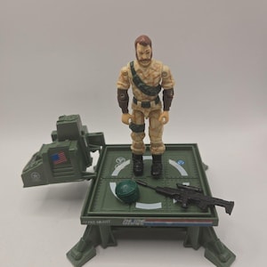 May include: A G.I. Joe action figure with brown hair and a mustache, wearing a tan camouflage uniform. The figure stands on a green plastic platform with a small green helmet and a black rifle. The platform features the text GIJOE and PAD ADJUST.