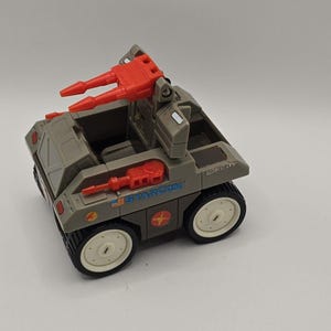 May include: A grey plastic Starcom toy vehicle featuring white wheels, orange dual cannons mounted on a folding frame, and an orange side-mounted weapon. The side of the vehicle displays the Starcom logo, a small American flag, and circular decals.