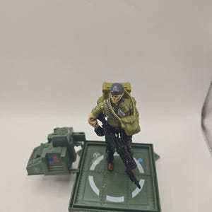 May include: A G.I. Joe action figure wearing a green shirt, black pants, and a dark blue headband, holding a black rifle. The figure stands on a green square platform with a white circular marking, featuring an attached green equipment console.