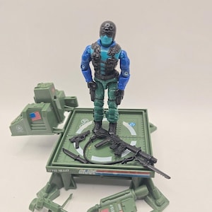 May include: A G.I. Joe action figure with a blue mask and black helmet, wearing a blue and green uniform with a black vest. The figure stands on a green platform with a rifle, pistol, and knife. Accessories include a green backpack with a U.S. flag.