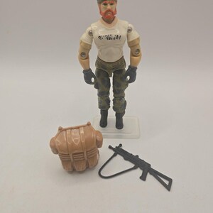 May include: An action figure with orange hair and a beard, wearing a white t-shirt with the word SURVIVAL printed on the chest, camouflage pants, and black gloves. Included are a tan plastic backpack accessory and a black toy rifle.