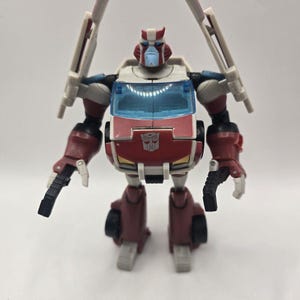 May include: A red and white robot action figure with a light blue face and translucent blue windscreen chest piece. The figure features black joints, two black handheld accessories, and two long white vertical components extending from the shoulders.