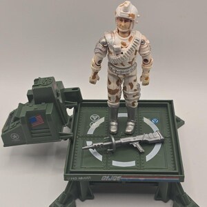 May include: A G.I. Joe action figure wearing a white and tan camouflage uniform and helmet, standing on a green plastic platform with a control console. A silver rifle lies on the platform, which features the text PAD ADJUST and G.I. JOE.
