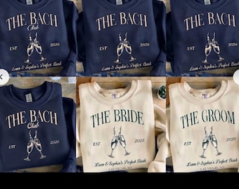 May include: A collection of navy blue and cream crewneck sweatshirts featuring the text THE BACH CLUB, THE BRIDE, or THE GROOM, with a graphic of two clinking champagne glasses, the year 2026, and personalized names printed on the front.