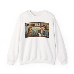 Könnte beinhalten: Ein weißes Sweatshirt mit Rundhalsausschnitt und einem Grafikdruck einer Büroszene mit dem Wort INTERVIEW oben. Die Illustration zeigt zwei Personen, die sich an einem Schreibtisch gegenübersitzen, mit einer Sprechblase mit Text.
