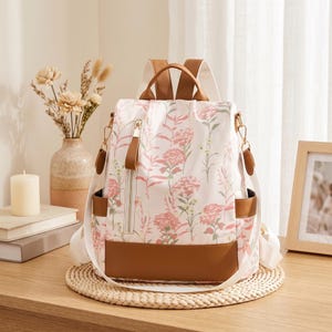 May include: A white backpack featuring a pink and green floral botanical print, accented with brown faux leather trim on the base, side pockets, and top handle. The bag includes a vertical zippered front pocket with a brown pull tab.