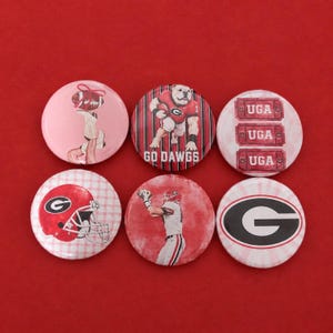 May include: Six circular pinback buttons featuring University of Georgia themes. Designs include a bulldog mascot, a football player, a red helmet with a G logo, three UGA tickets, a large G logo, and a stylized illustration of a person with a football.
