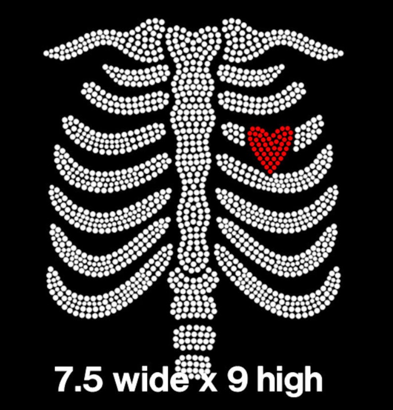 May include: White rhinestone design of a ribcage with a red rhinestone heart in the center. 7.5 inches wide by 9 inches high.