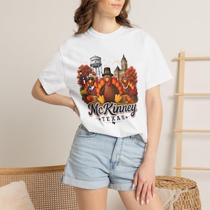Peut inclure: Un t-shirt blanc à manches courtes avec un motif représentant trois dindes devant un château d'eau et un bâtiment, avec le texte McKinney Texas. Les dindes portent des chapeaux et des écharpes, entourées de feuillage d'automne et de citrouilles.
