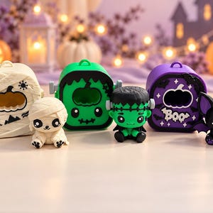May include: Three small Halloween-themed cases and three matching figurines. One cream mummy case with a mummy figure, one green Frankenstein case with a Frankenstein figure, and one purple bat case with a black bat figure. Each case has a pumpkin cutout.