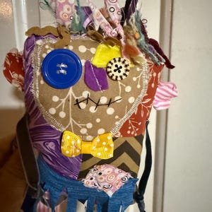 May include: A handcrafted fabric doll constructed from various patterned textile scraps. The face features a large blue button eye, a smaller patterned button eye, a yellow bow tie, and stitched mouth details. The head is adorned with frayed fabric strips.