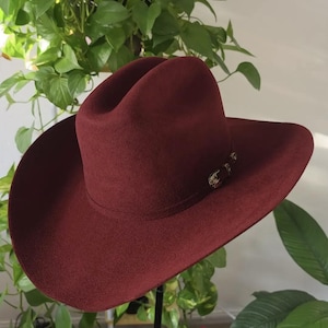 Op de afbeelding: Een diepbordeauxrode vilten cowboyhoed met een gestructureerde kroon en een brede, gebogen rand. De hoed is voorzien van een bijpassende hoedband die aan de zijkant is versierd met een kleine, zilverkleurige metalen gesp als subtiel accent.