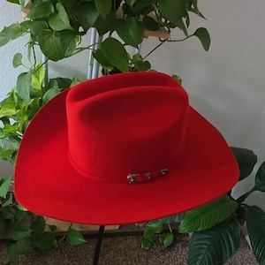 Op de afbeelding: Een felrode cowboyhoed met een gestructureerde kroon met een centrale vouw en een brede, gebogen rand. Een dunne, bijpassende rode hoedband omcirkelt de basis van de kroon, geaccentueerd met een klein metalen gespdetail aan de voorzijde.