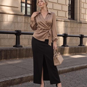 May include: A satin wrap blouse in a muted tan color with long sleeves and a side tie, paired with a floor-length black skirt featuring a central front slit, black pointed-toe high heels, and a small beige structured handbag.