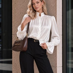 May include: A white satin long-sleeve blouse with a high collar, button-down front, and flared cuffs, paired with black trousers featuring a thin black belt and a dark brown leather shoulder bag with a gold-toned metal horsebit detail.
