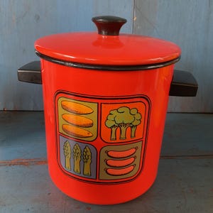 May include: A vintage red enamel cooking pot with a lid and black handles. The pot features a decorative panel with illustrations of vegetables, including corn, broccoli, asparagus, and sausages. The pot is a classic kitchenware item.