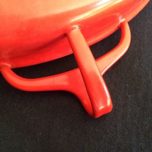 May include: A bright orange enameled cast iron cookware piece featuring a smooth, glossy finish and a curved, integrated side handle. The design includes a sturdy, rounded loop handle attached to the main body, showcasing a vibrant, solid color.