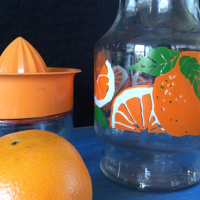 Pu&ograve; includere: Un set di utensili da cucina a tema arancia, tra cui una caraffa di vetro con fette di arancia stampate, uno spremiagrumi di plastica e un'arancia intera.