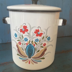 May include: A white enamel stockpot with a black rim and black side handles. The lid features a black knob. The front is decorated with a colorful floral pattern in red, blue, and brown, featuring stylized leaves and a central blue oval motif.