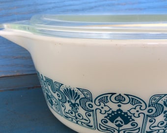 May include: White casserole dish with a clear glass lid. The dish features a teal floral pattern around the base. It has a handle on one side. The dish is made of a thick, durable material.