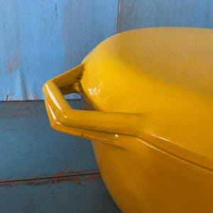 May include: Close-up of a bright yellow, oval-shaped cooking pot with a matching lid. The pot has two sturdy handles and a glossy finish. The background is a mix of blue and turquoise painted wood.