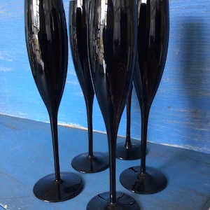 May include: Four black champagne flutes with a tall, slender stem and a wide bowl. The flutes are arranged in a row on a blue surface.