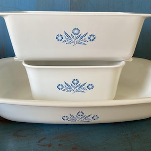 May include: A set of three white rectangular baking dishes of varying sizes stacked vertically. Each dish features a centred blue floral pattern consisting of three stylised flowers with leaves. The dishes have smooth, rounded edges and a glossy finish.