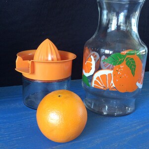 Pu&ograve; includere: Set da cucina a tema arancia con una caraffa di vetro con fette di arancia e uno spremiagrumi in vetro con parte superiore arancione.