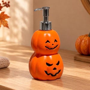 Può includere: Un dispenser di sapone in ceramica arancione brillante a forma di due zucche sovrapposte. La zucca superiore ha un volto sorridente, mentre quella inferiore presenta un ghigno con denti aguzzi. Include una pompa in metallo color argento.