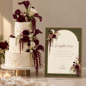 May include: A three-tiered white cake decorated with dark red calla lilies, deep red flowers, and blackberries, displayed next to a framed sign that reads Tonights tiers, Pistachio & Blackberry, Matcha & White Chocolate Raspberry, Lemon, Olive Oil & Rosemary, Liam and Scarlett.
