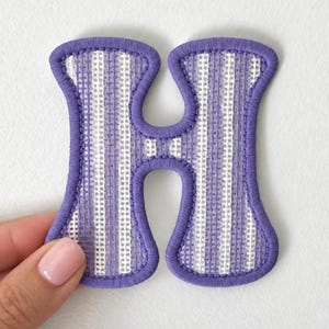 May include: A letter H patch featuring a purple and white vertical striped pattern with a textured fabric weave. The letter is outlined with a thick, raised purple embroidered border that defines its shape and provides a clean edge.