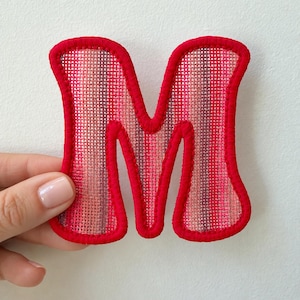 May include: A letter M patch featuring a woven fabric texture with vertical stripes in shades of pink, red, and purple. The letter is outlined with a thick, raised red embroidered border that defines its shape against the background.
