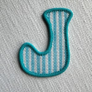 May include: An embroidered letter J patch featuring a vertical striped pattern in white and light blue. The letter is outlined with a thick, solid teal border and has a textured, woven fabric appearance throughout the entire shape.