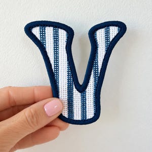 May include: A fabric letter V patch featuring a white and navy blue vertical striped pattern. The letter has a thick, dark navy blue stitched border that outlines the shape, creating a clean and defined edge for the textile applique.