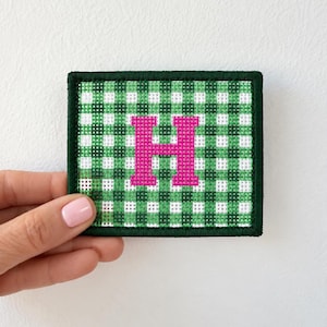 May include: A rectangular embroidered patch featuring a green and white gingham checkered pattern. Centered on the patch is a bold, bright pink capital letter H. The entire patch is finished with a dark green stitched border edge.