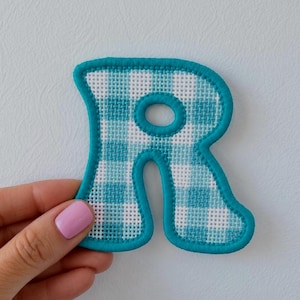 May include: A fabric letter R patch featuring a light blue and white gingham check pattern. The letter has a thick, solid teal blue embroidered border that outlines the entire shape, providing a distinct edge to the checkered fabric.