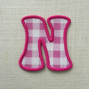 May include: A fabric letter N patch featuring a pink and white gingham check pattern. The letter has a thick, solid pink embroidered border that outlines the shape, creating a distinct edge against the textured, woven fabric background.