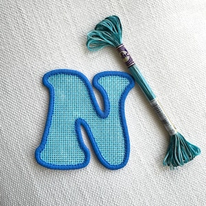 May include: A light blue letter N patch with a textured weave pattern and a dark blue stitched border, accompanied by a skein of variegated teal and blue embroidery floss with a label marked DMC and the number 025.