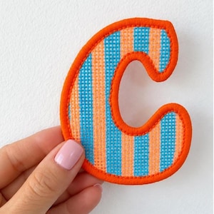 May include: A letter C patch featuring a woven fabric texture with vertical stripes in alternating shades of bright orange and light blue. The entire edge of the letter is finished with a thick, vibrant orange embroidered border.