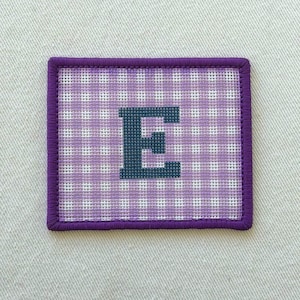 May include: A rectangular needlepoint patch featuring a light purple and white gingham check pattern. The letter E is stitched in the center in a dark blue thread. The entire patch is bordered by a thick, dark purple stitched edge.