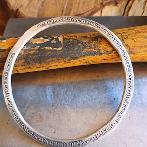 Puede incluir: Un brazalete de plata con la oración del Señor grabada alrededor del exterior. El brazalete está sobre una superficie de madera.