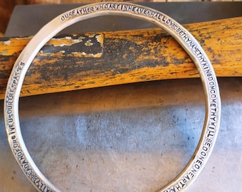 May include: A silver bangle bracelet with the Lord's Prayer engraved around the outside. The bracelet is on a wooden surface.