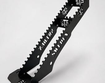 May include: A black metal stand with a serrated top edge and a flat base. The structure features cut-out text reading SWEDEN and the letters STP. The design includes two parallel vertical supports connected by crossbars, with circular holes.