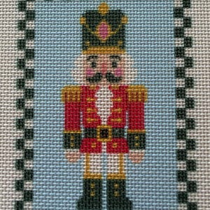 May include: A needlepoint canvas featuring a nutcracker soldier in a red jacket, gold epaulets, and a black hat with a pink gem. The figure is centered on a light blue background, enclosed by a black and white checkered border pattern.