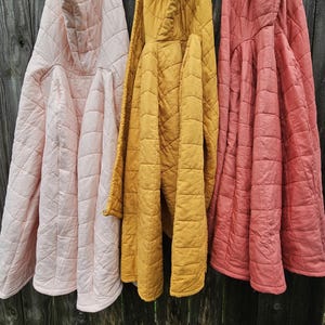 May include: Three hooded jackets in light pink, mustard yellow, and coral red. Each jacket features a quilted diamond pattern across the fabric, long sleeves, and a hood. The garments are displayed hanging side by side in a vertical orientation.