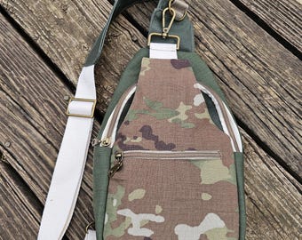 May include: A green and camouflage sling bag with a white strap and gold-colored hardware. The bag features a front zipper pocket and a main compartment with a zipper closure. The bag is made of durable fabric.