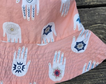 May include: A peach-colored bucket hat featuring a repeating pattern of white hand illustrations. Each hand design includes intricate blue and red geometric symbols, stars, and eye motifs on the palms and fingers, with visible stitching along the brim.